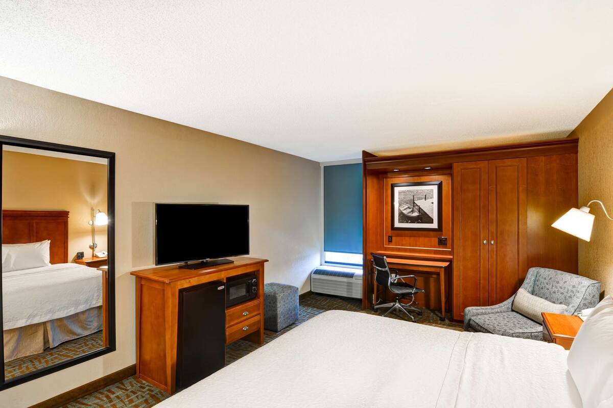 Ein Hotelzimmer mit einem großen Bett, einem Schreibtisch mit Flachbildfernseher, einem Sessel, einer Schreibtischlampe und einem gerahmten Bild an der Wand.