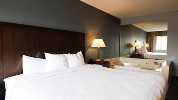 Quarto de hotel com uma cama grande, uma mesa de cabeceira com abajur, e banheiro com pia e espelho.