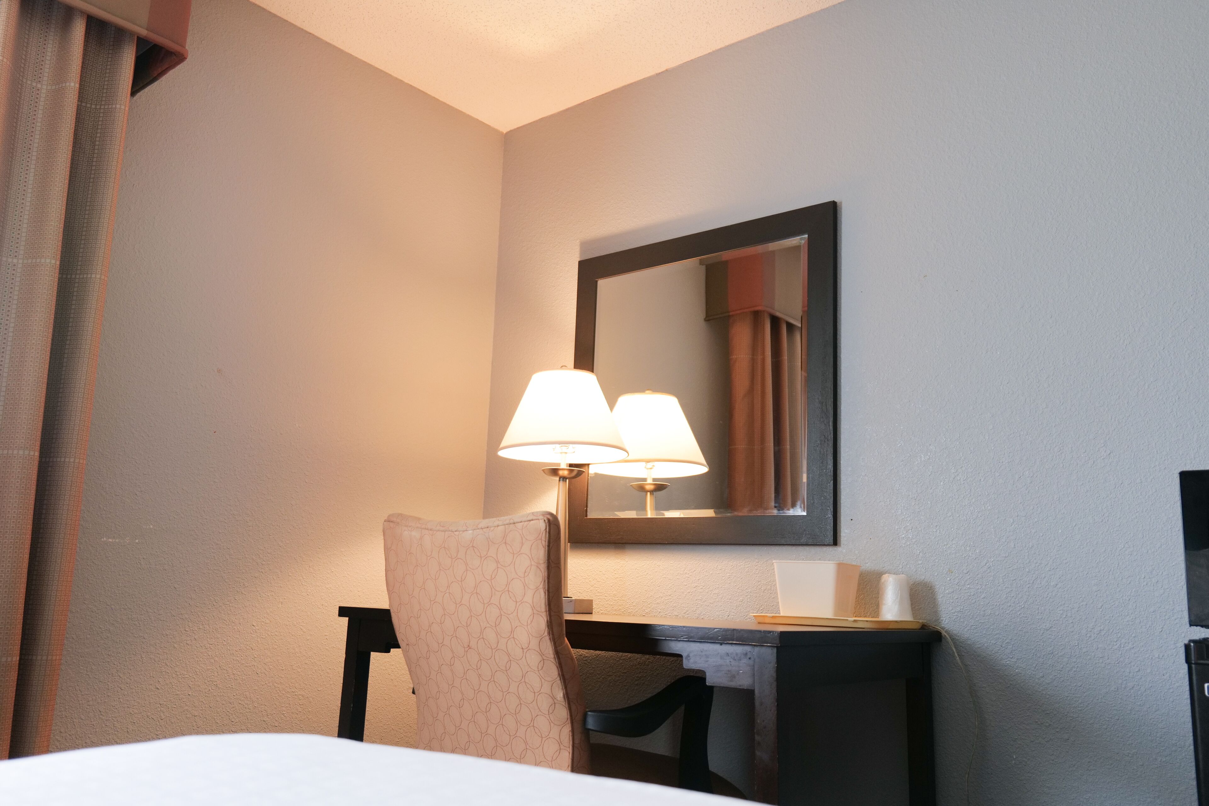 Habitación de hotel con una cama, un escritorio con lámpara, una silla y un espejo en la pared.