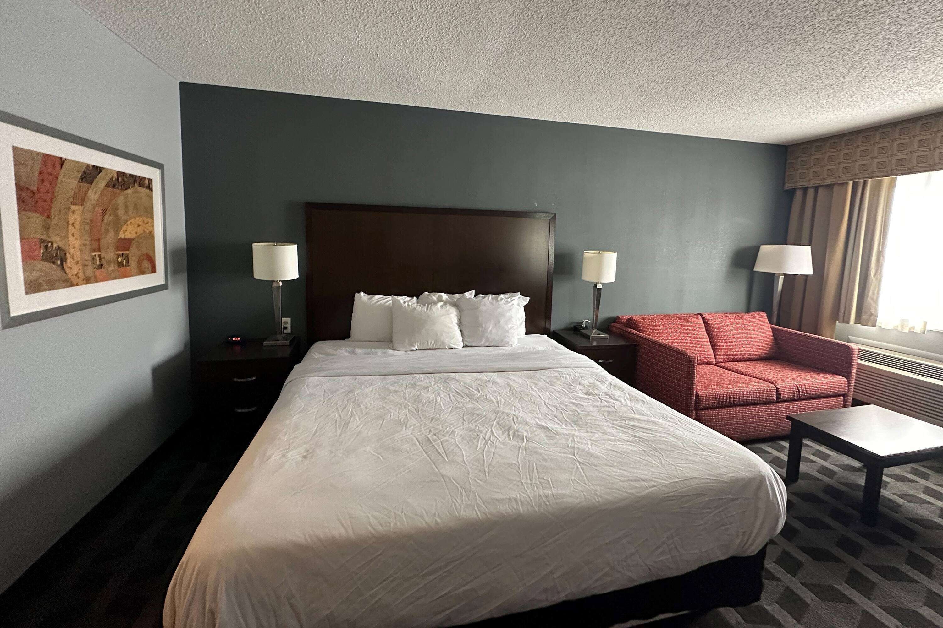 Ein Hotelzimmer mit einem großen Bett, zwei Nachttischen, einem roten Sessel, einem kleinen Tisch und einem Bild an der Wand.