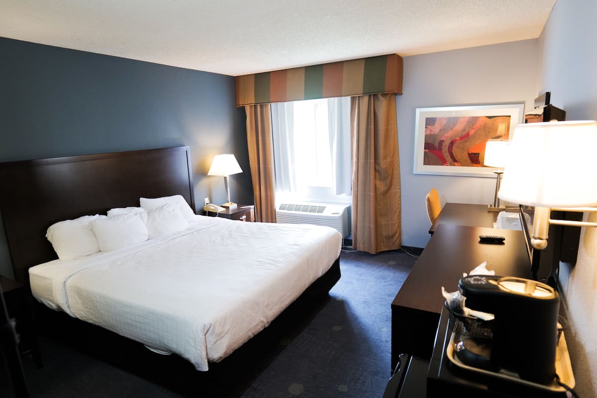 A hotel room with a large bed, a desk, a chair, a lamp, an air conditioner, and a painting on the wall.