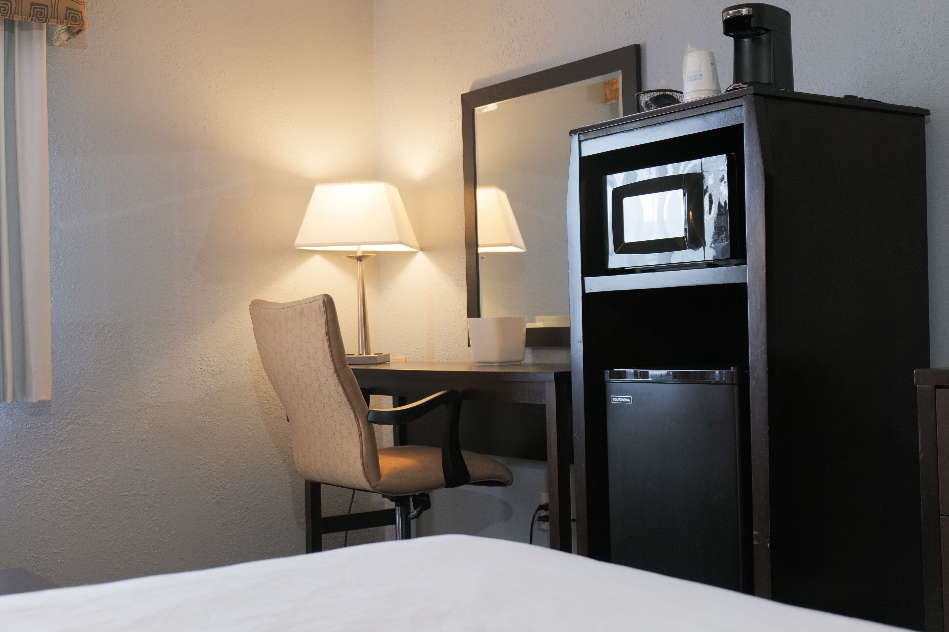 A hotel room with a bed, a desk with a chair, a lamp, a refrigerator, and a mirror.