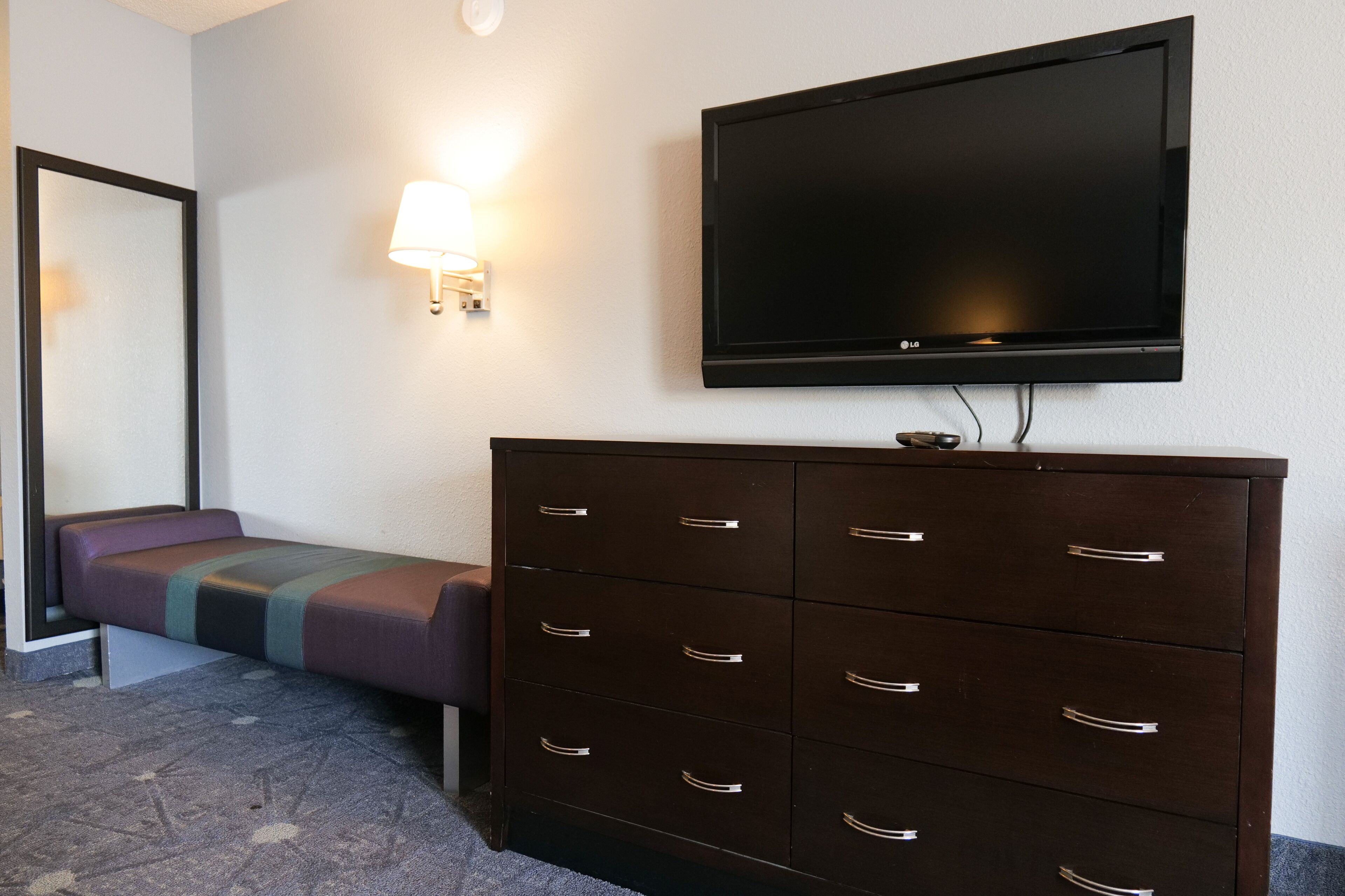 A hotel room with a bed, a dresser, a television, and a wall-mounted lamp.