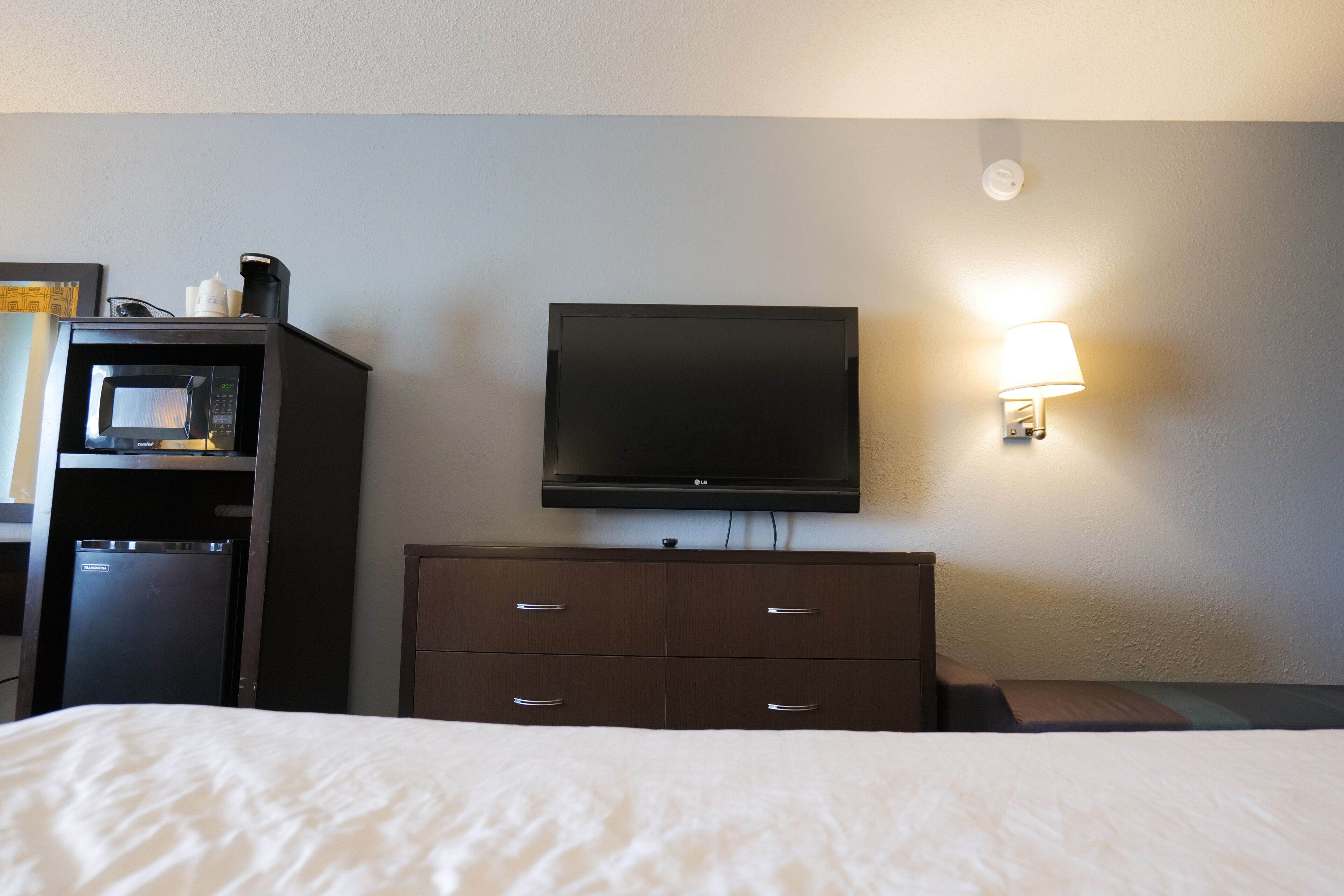 A hotel room with a bed, a television mounted on the wall, a small refrigerator, and a wall-mounted lamp.