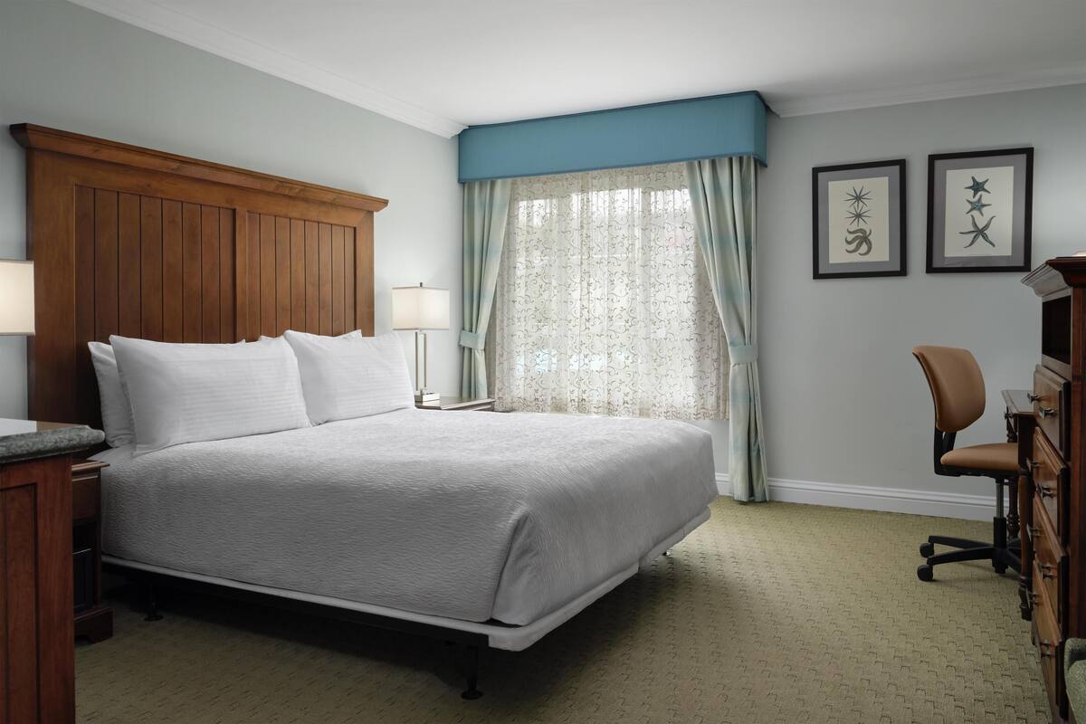A neatly arranged bedroom with a large bed, a wooden headboard, a desk with a chair, and two framed pictures on the wall.