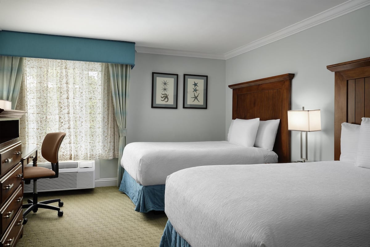 A hotel room with two beds, a desk with a chair, a lamp, and framed artwork on the wall.