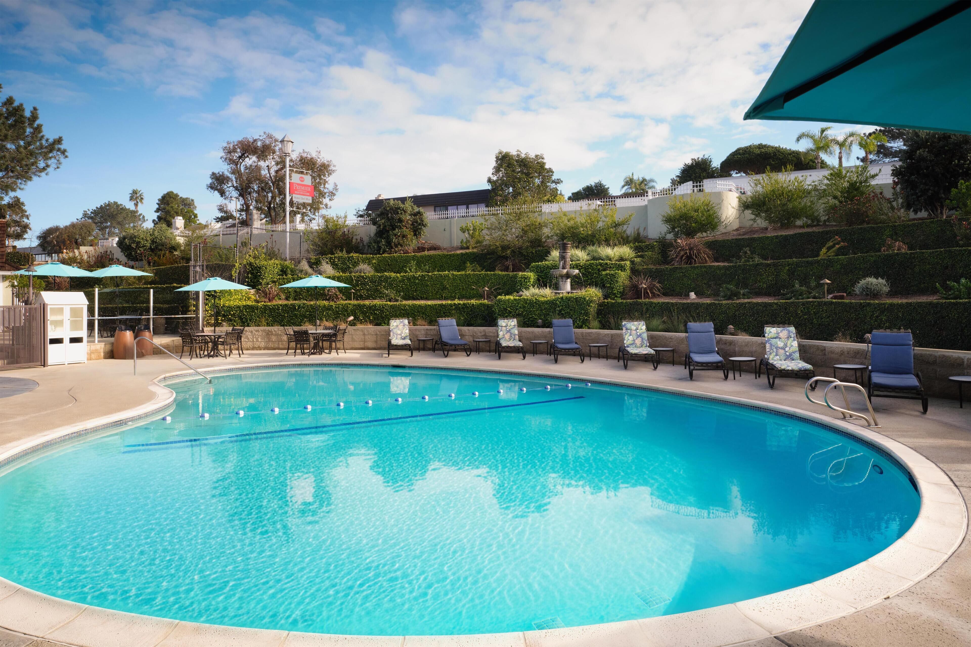 A swimming pool with lounge chairs and umbrellas, surrounded by landscaped gardens and a building in the background.