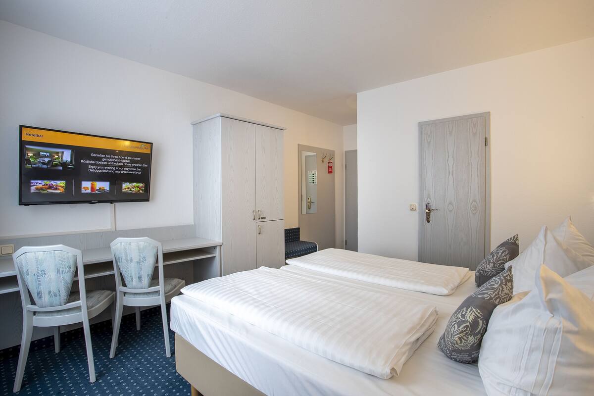A hotel room with a bed, two chairs, a desk, and a TV displaying a menu.