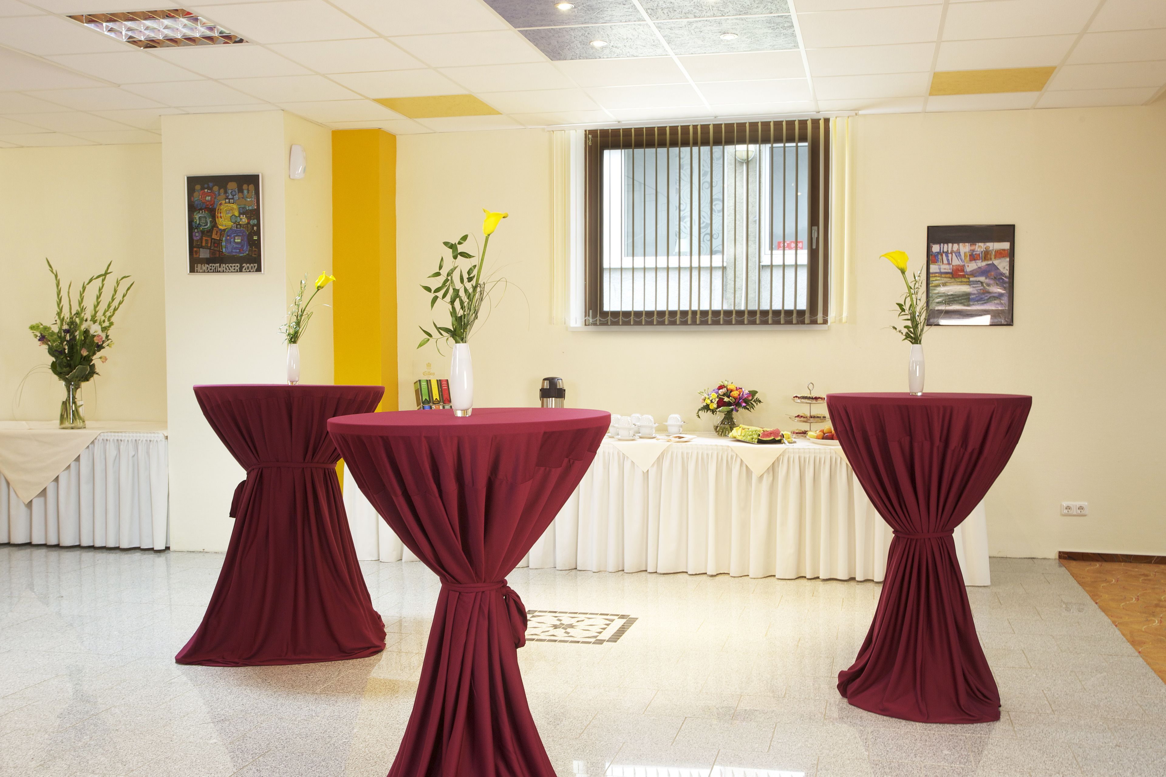 Een vergaderzaal met drie tafels, bedekt met bordeauxrode tafelkleden, witte tafellinnen en bloemstukken als decoratie.