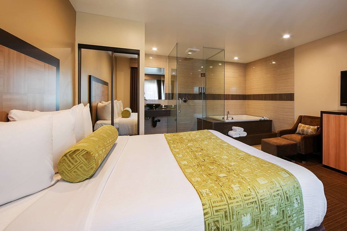 Une chambre d’hôtel dotée d’un grand lit, d’un coin salon avec un fauteuil et un pouf, d’une télévision, d’une salle de bain avec douche et d’un miroir.