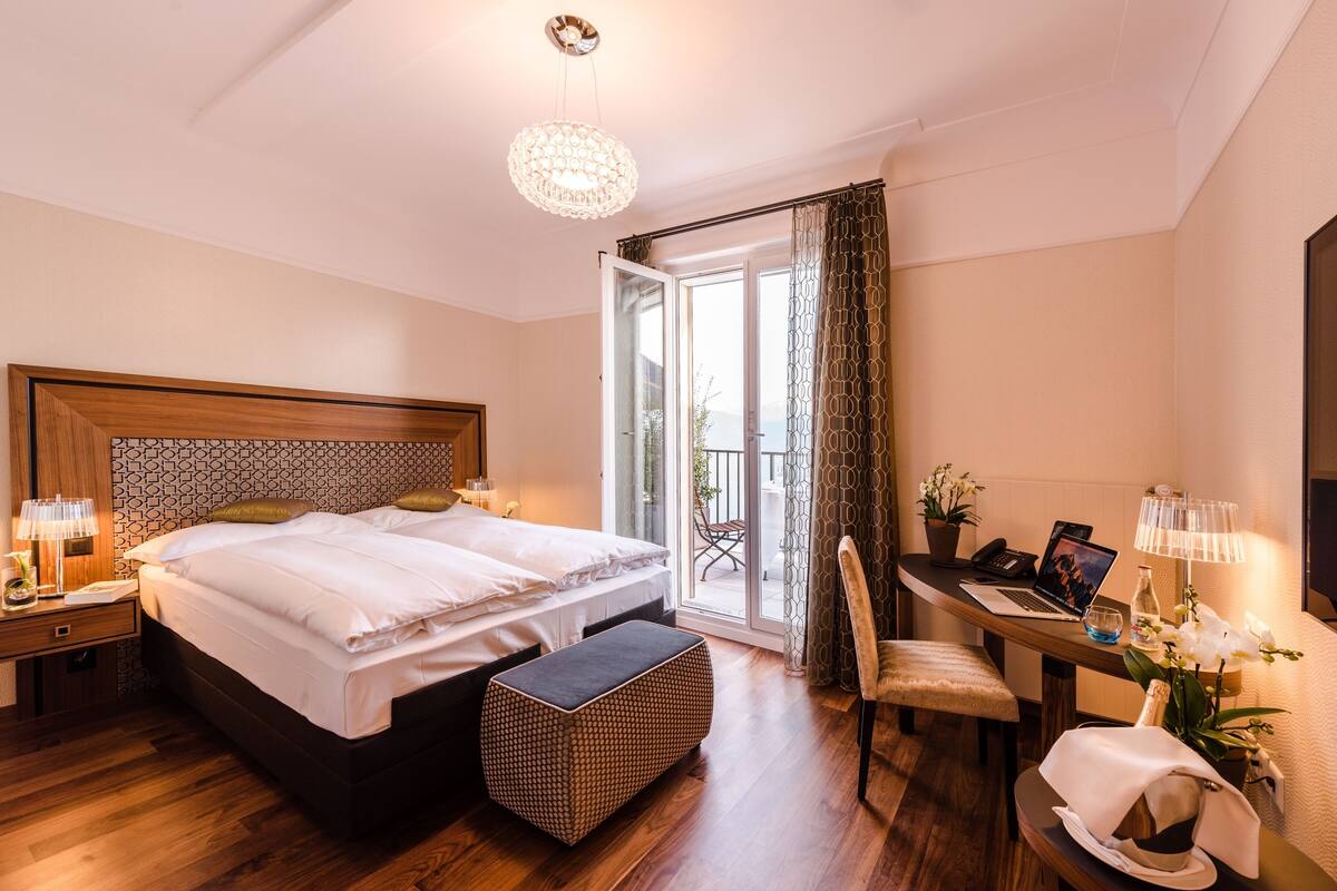 Een hotelkamer met een groot bed, een bureau met een laptop, een stoel en een balkon met gordijnen.