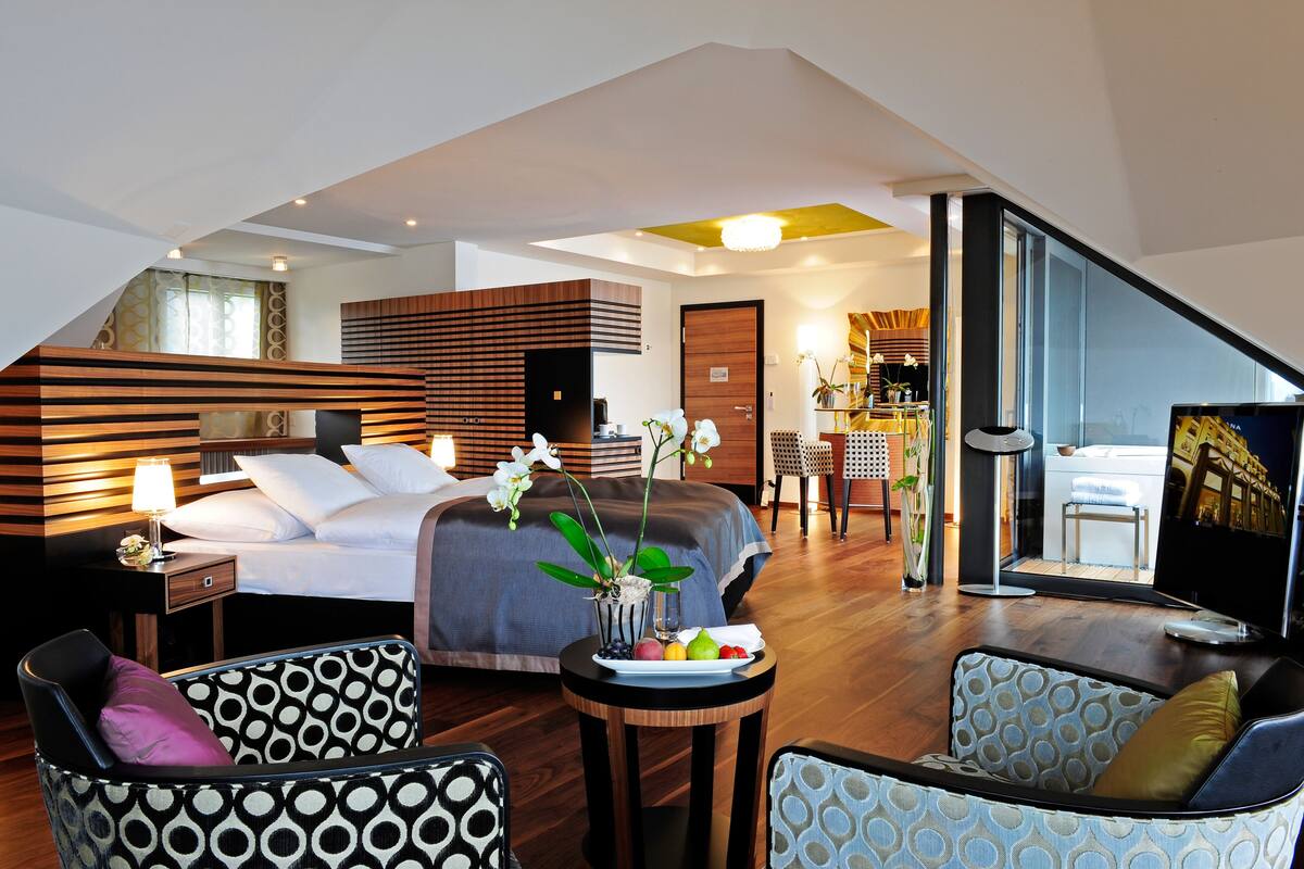 Een moderne hotelkamer met een groot bed, een zithoek met gestreepte stoelen, een kleine tafel met fruit en een televisie.