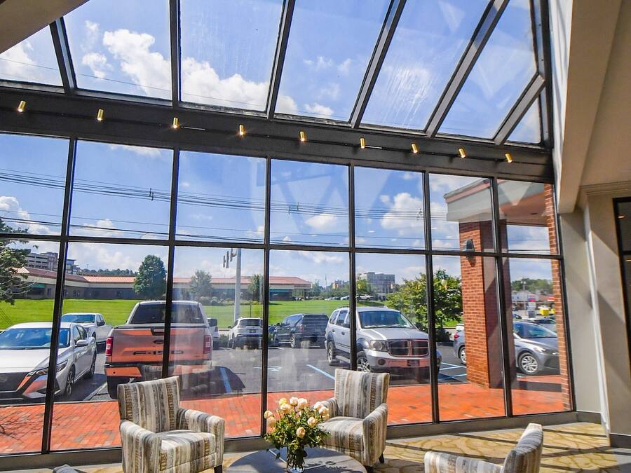 A spacious indoor area with large windows overlooking a parking lot. There are several upholstered chairs arranged around a central table with a vase of flowers.
