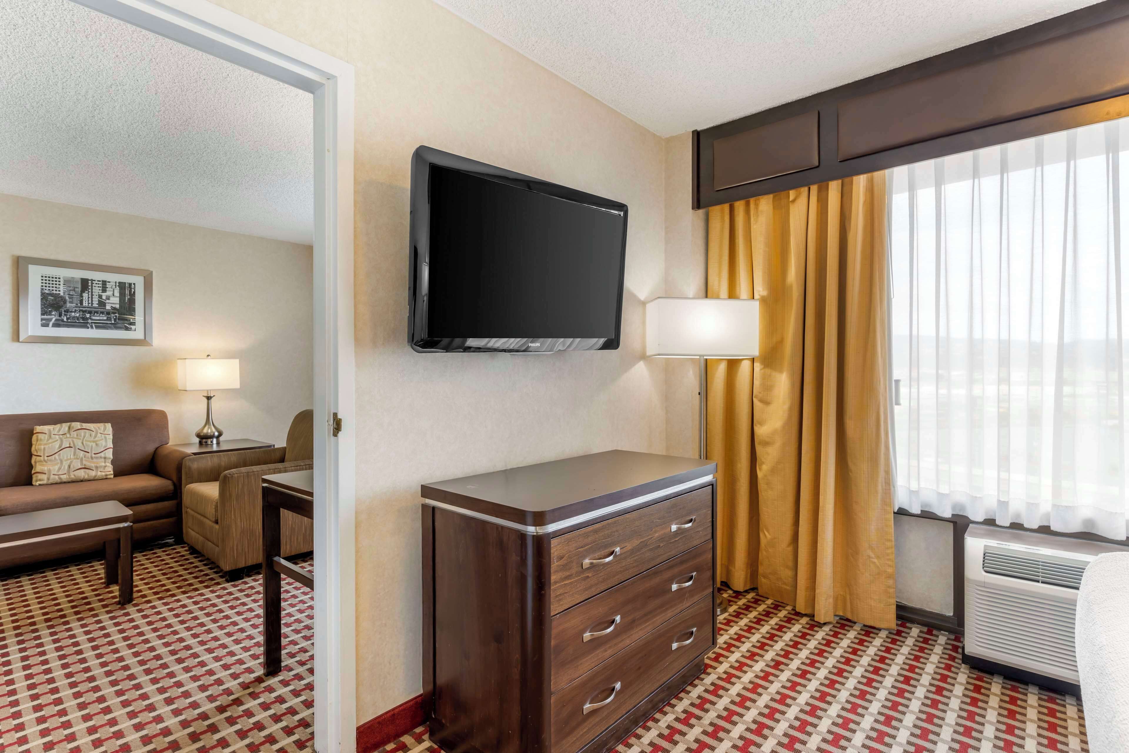 Habitación de hotel con una televisión de pantalla plana montada en la pared, un escritorio con lámpara, un sofá, un aparador y una ventana con cortinas.