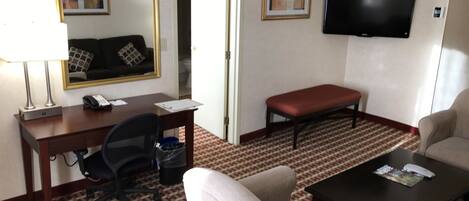 A hotel room with a desk, office chair, two armchairs, a red bench, a television, and a patterned carpet.