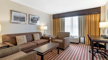 A hotel room with a brown sofa, armchairs, a small table, a desk, and a patterned carpet. There are two framed pictures on the wall, a lamp, and a window with curtains.
