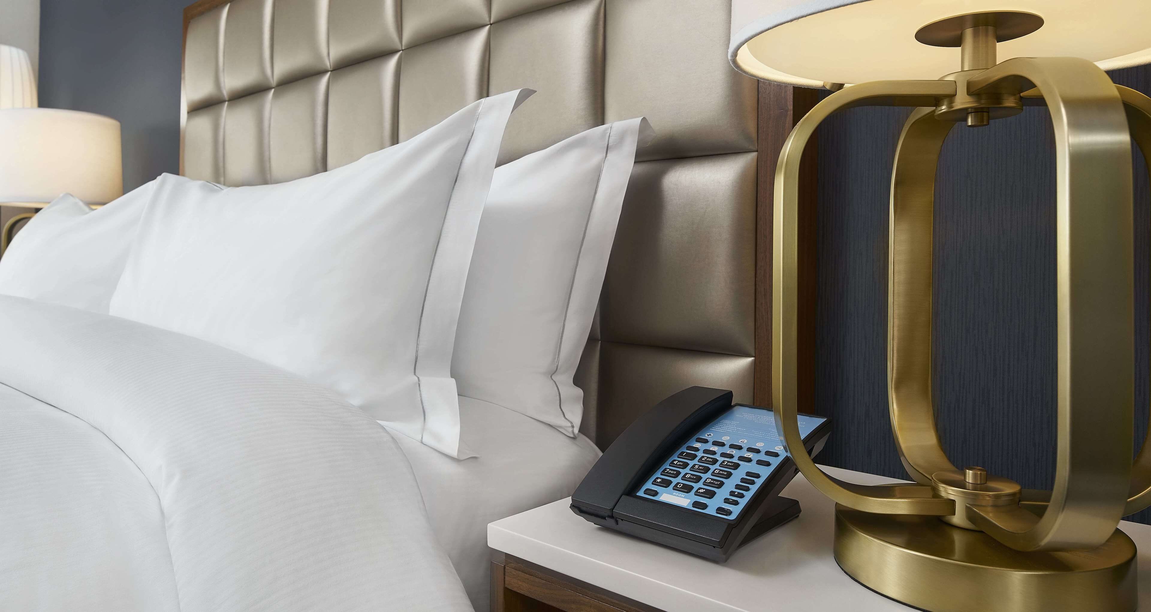 Et hotelværelse med en polstret sengegavl, en telefon ved siden af sengen og en lampe med gylden ramme på natbordet.