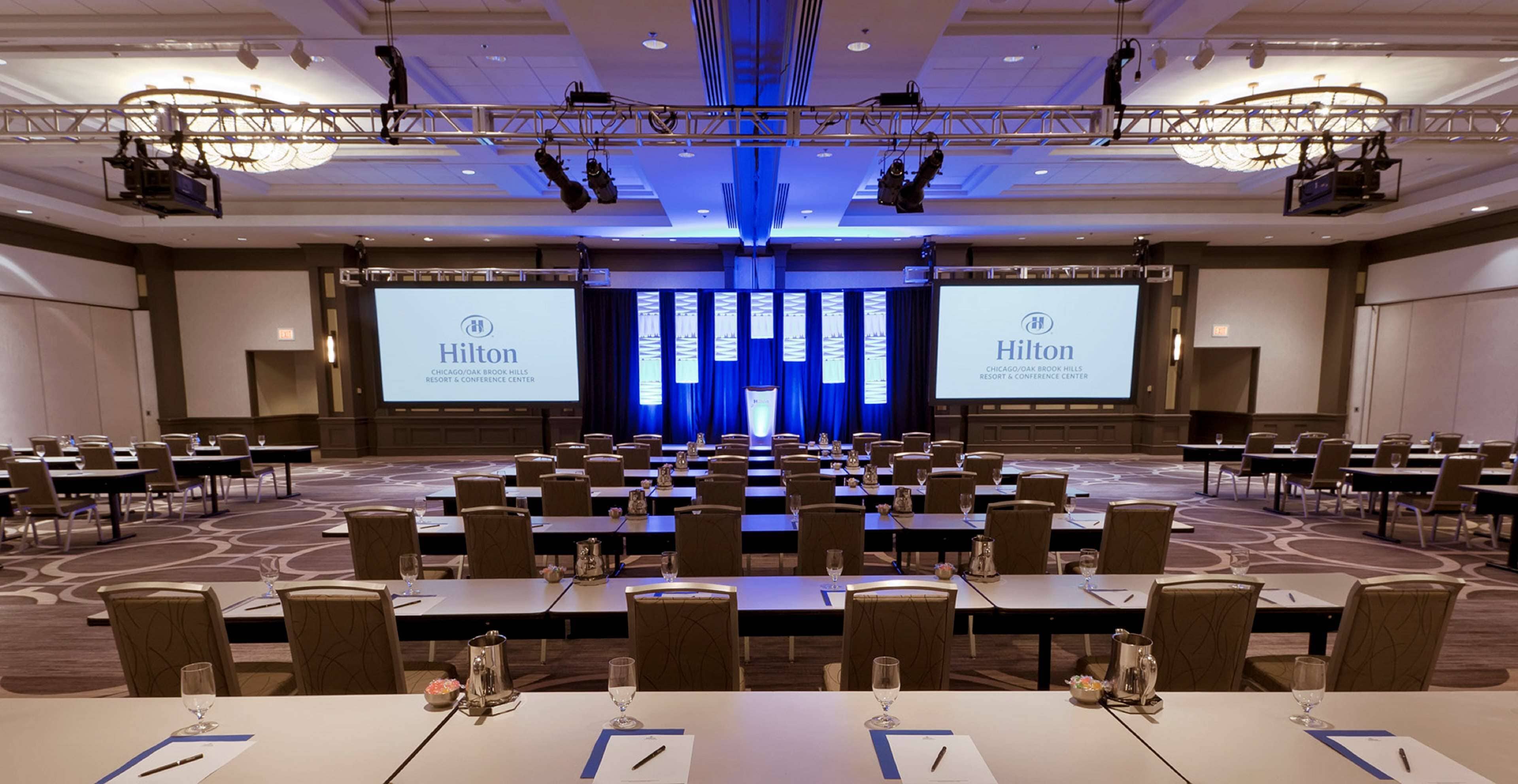 A large conference room with rows of tables and chairs, a stage with a blue backdrop, and two large screens displaying the Hilton logo.