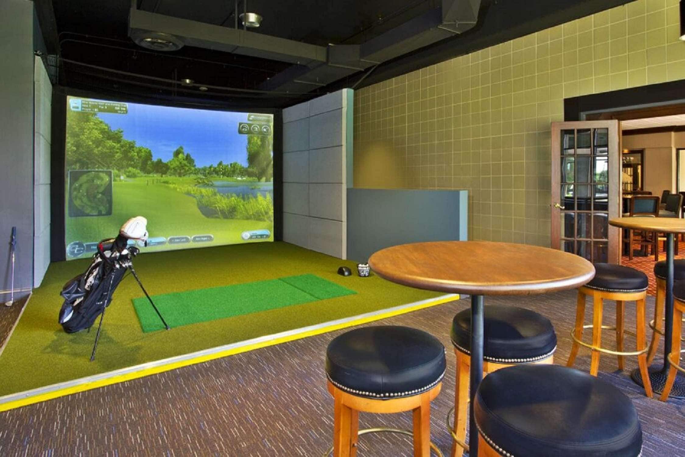 Una sala de simulación de golf con una pantalla grande, un bolso de golf y taburetes.