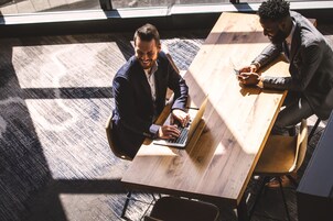 Two individuals seated at a wooden table with a laptop, in a room with a patterned carpet and large windows.