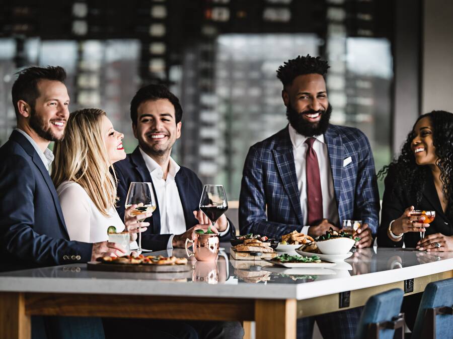 Un grupo de personas sentadas en una mesa con comida y bebidas, en un ambiente interior moderno con grandes ventanales.