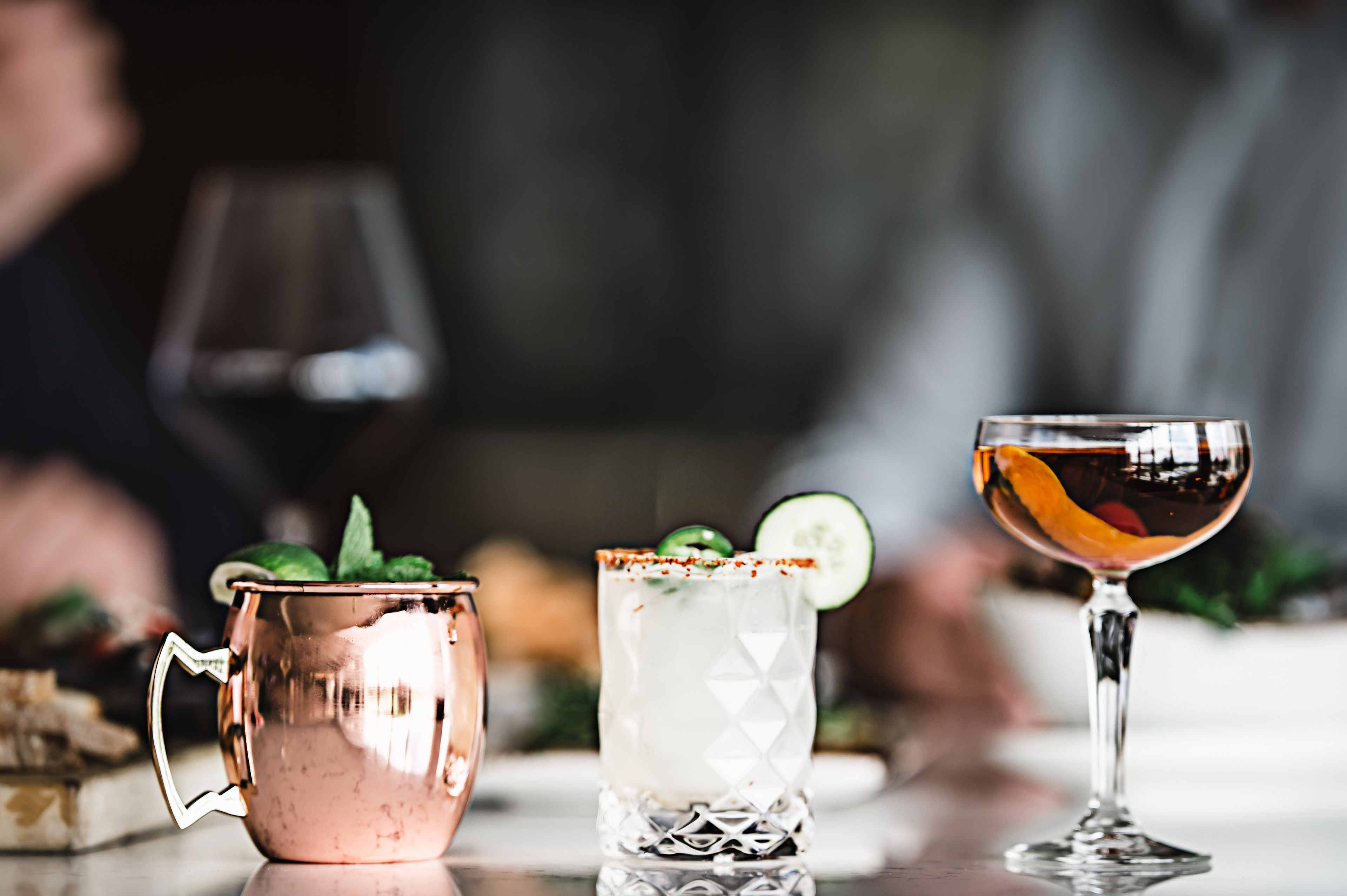 Trois boissons sont posées sur une table : une tasse en cuivre, un verre transparent avec une tranche de citron vert et un verre à martini avec une tranche d’orange.
