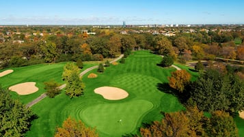 An aerial view of a golf course with sand bunkers, a putting green, and a city skyline in the background.