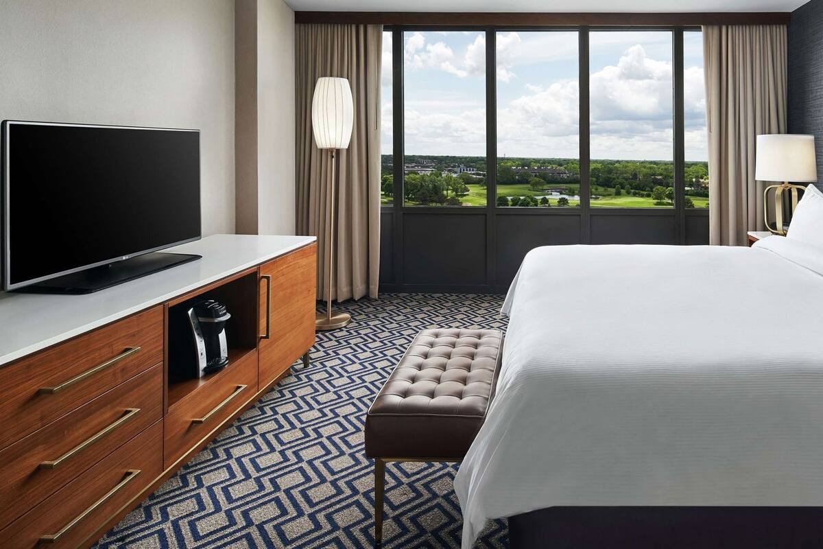 A hotel room with a large bed, a TV on a wooden cabinet, a view of a golf course, and a patterned floor.