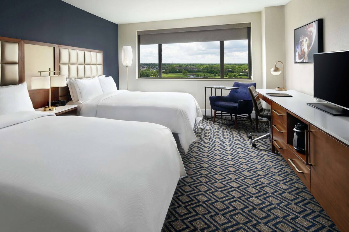 A hotel room with two beds, a desk, a chair, a TV, and a window with a view.