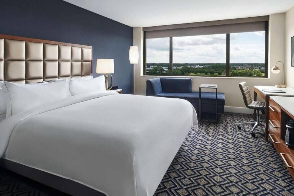A hotel room with a large bed, a desk, a chair, a TV, and a window with a city view.