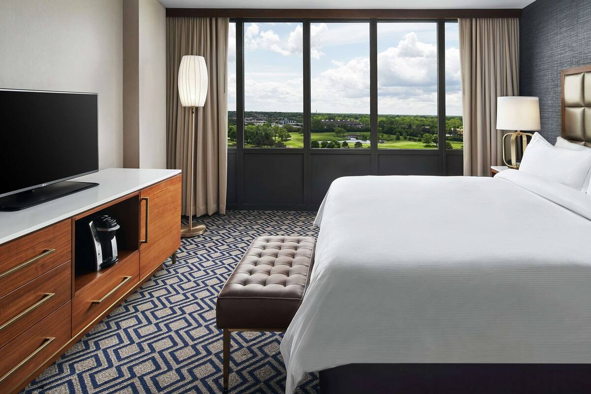 A hotel room with a large bed, a flat-screen TV, a wooden dresser, and a view of a green landscape through the window.