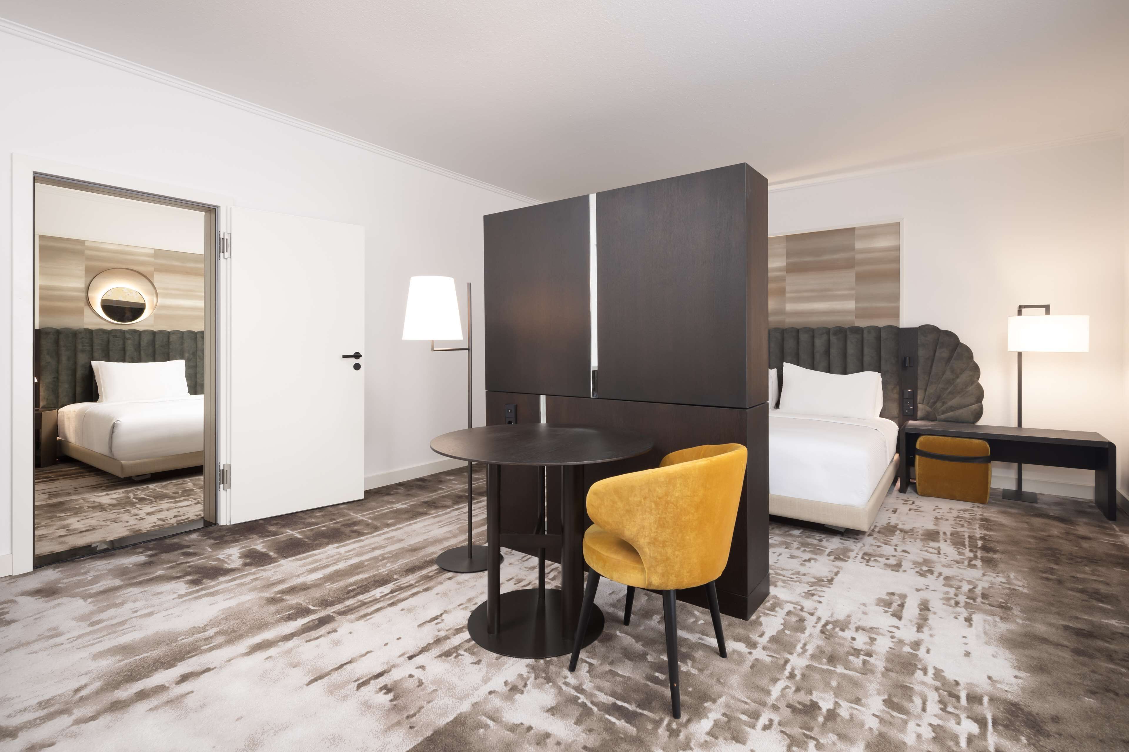 Une chambre d’hôtel avec un lit, une table ronde, une chaise jaune, une armoire de couleur sombre et une lampe.