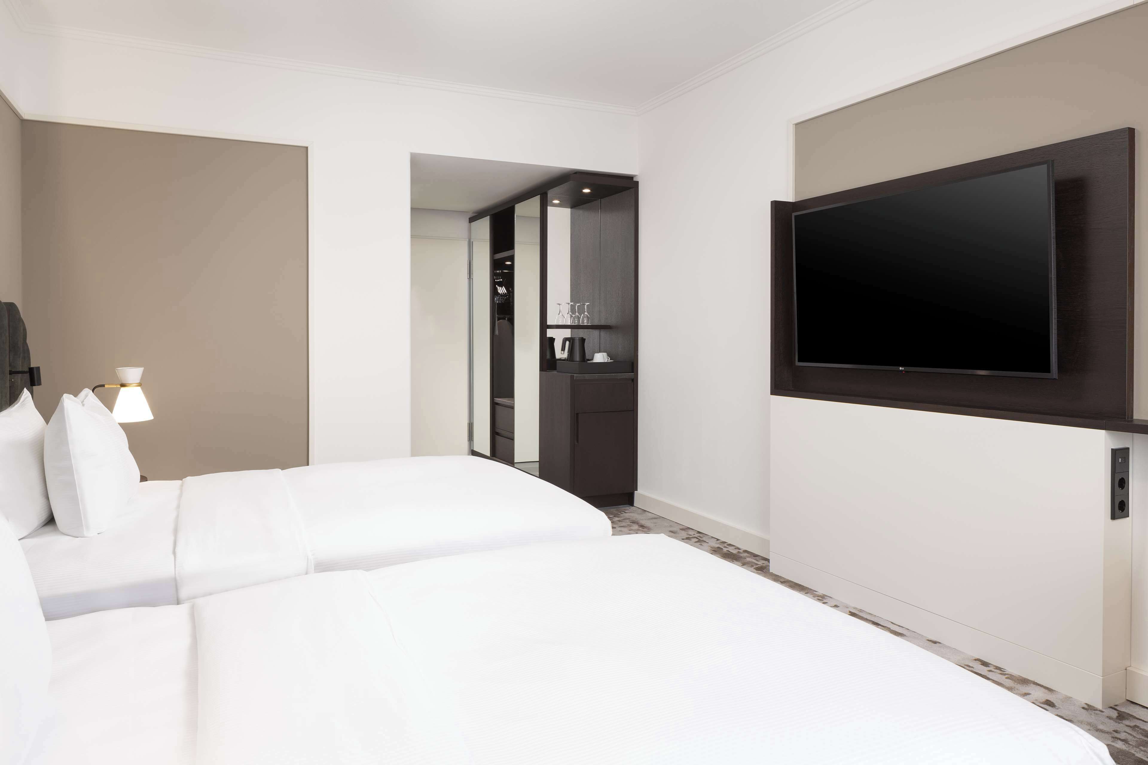 A hotel room with two beds, a flat-screen TV mounted on the wall, a walk-in closet, and a bathroom visible through an open door.