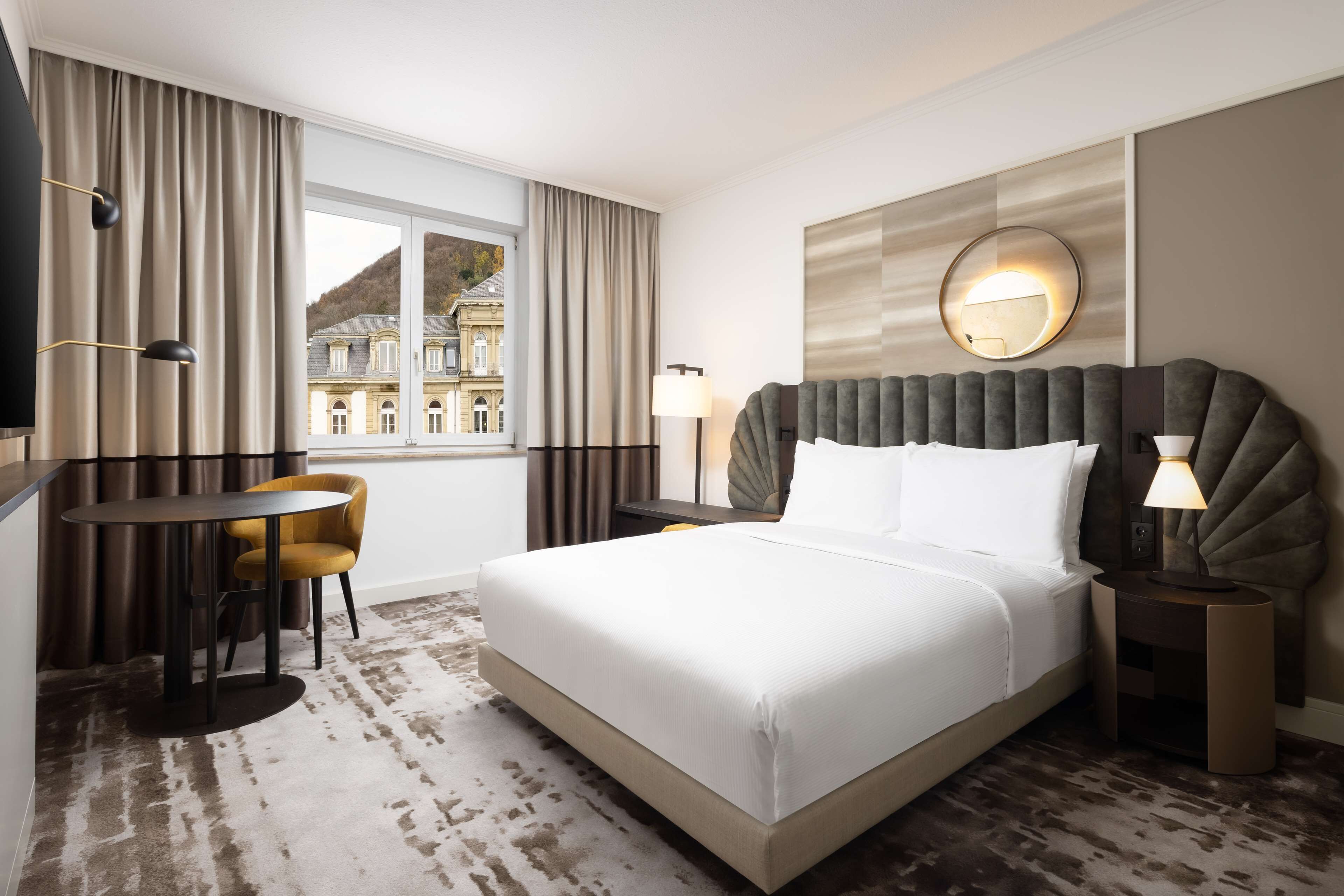 Habitación de hotel con una cama grande, un escritorio con silla, una mesa redonda y vistas a un edificio a través de una ventana.