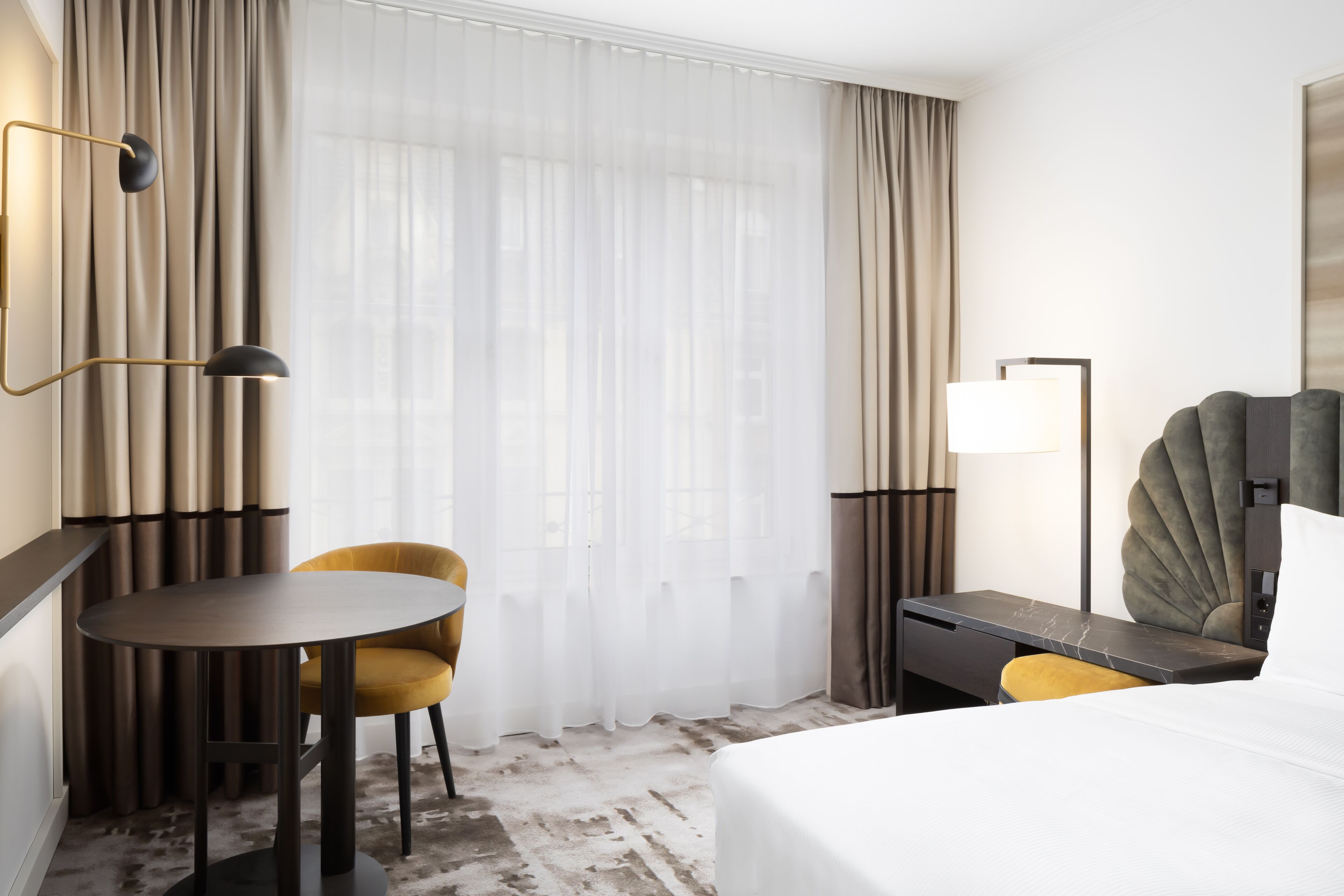 A hotel room with a bed, a desk, a chair, a lamp, and a large window with curtains.