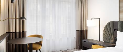 Habitación de hotel con cama, escritorio, silla, lámpara y una ventana grande con cortinas.