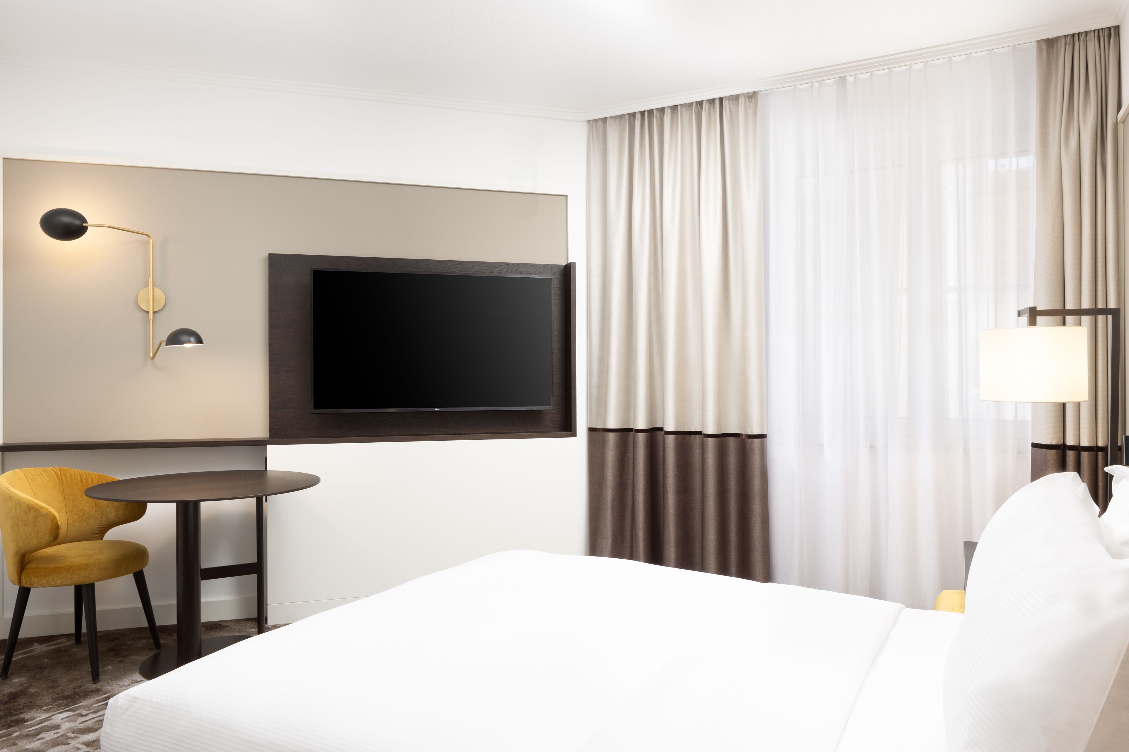 Une chambre d’hôtel avec un grand lit, un téléviseur à écran plat fixé au mur, une petite table ronde avec une chaise jaune et une lampe sur la table de chevet.
