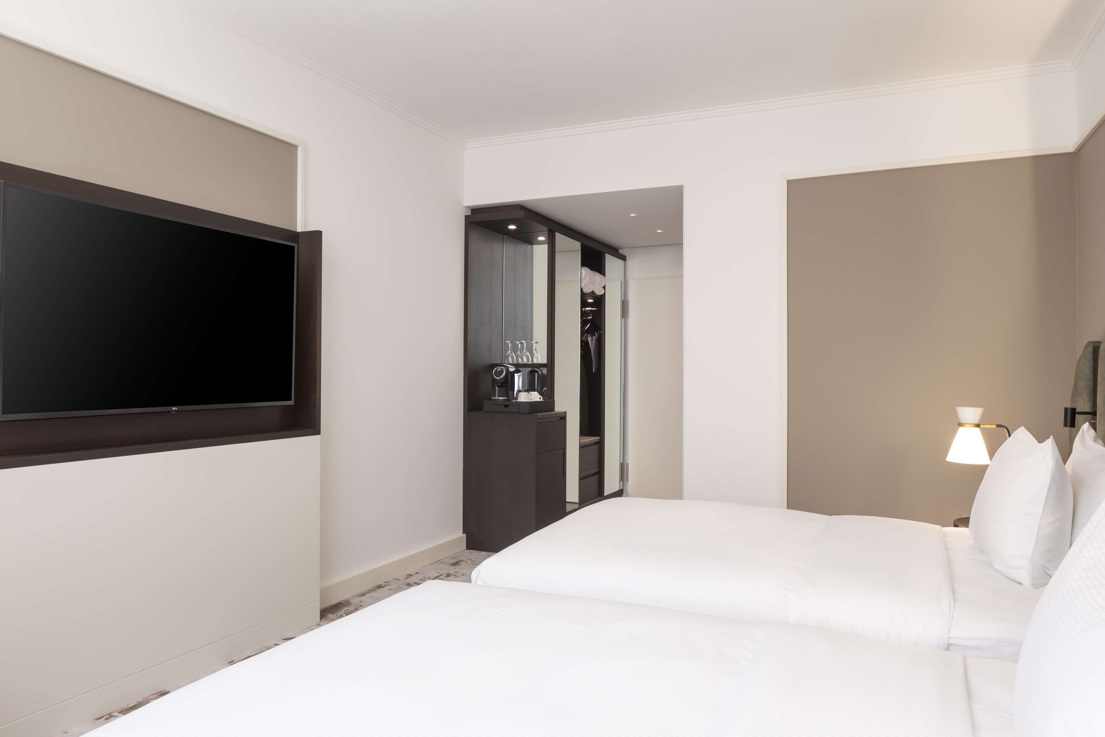 Quarto de hotel com televisão de tela plana grande fixada na parede, cama com roupa de cama branca, mesa de cabeceira com abajur e closet espaçoso com espelho e máquina de café.