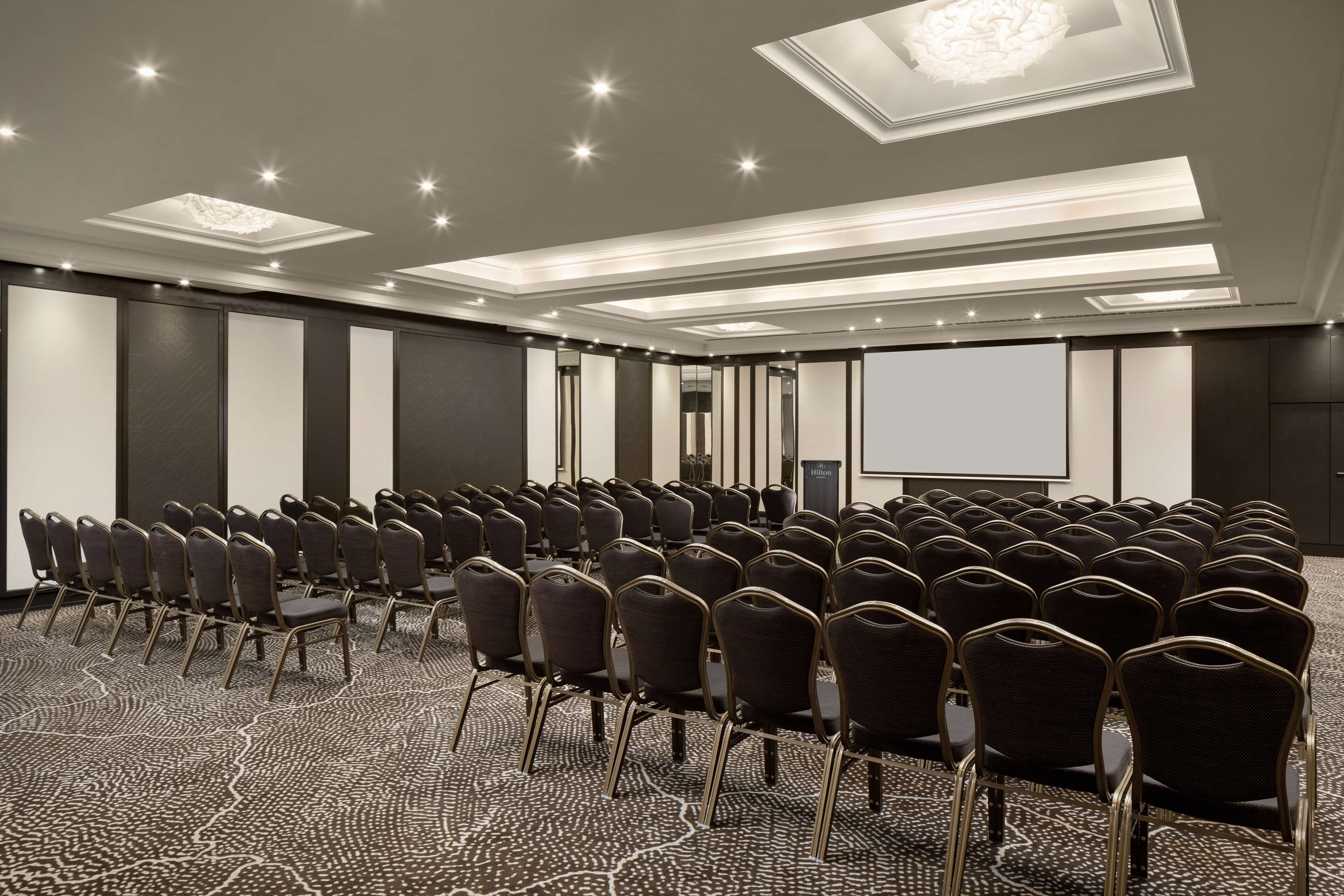 Uma sala de conferências com fileiras de cadeiras, uma tela de projetor e um tapete estampado.