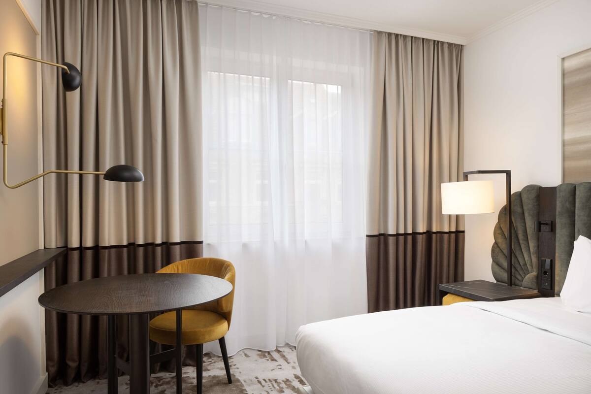 A hotel room with a bed, a round table, a chair, a desk with a lamp, and curtains.
