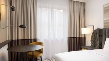 Ein Hotelzimmer mit einem Bett, einem runden Tisch, einem Sessel, einem Schreibtisch mit Lampe und Vorhängen.