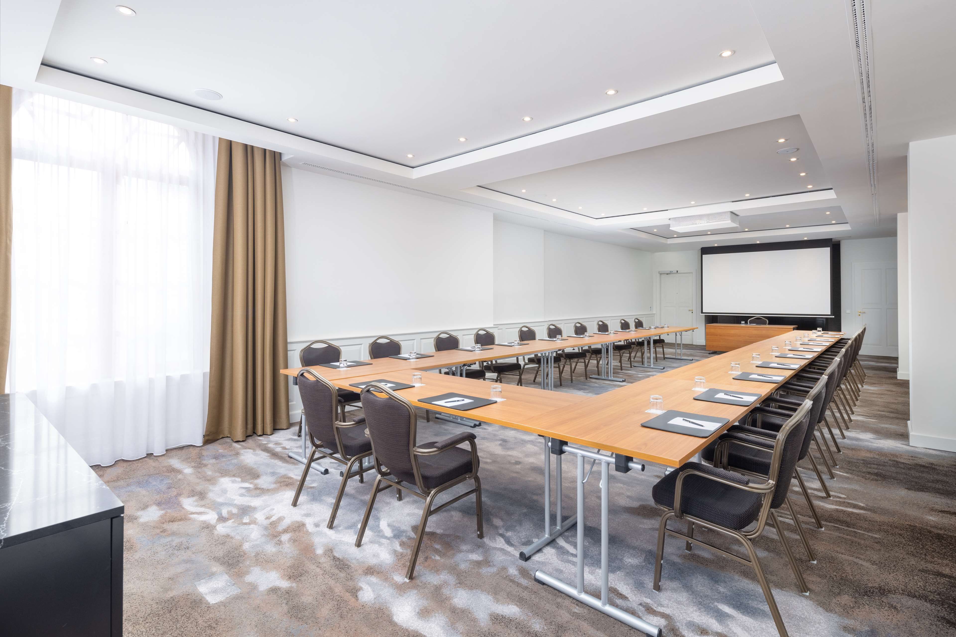 Une salle de conférence avec une longue table, des chaises, un écran de projection et une grande fenêtre avec des rideaux.