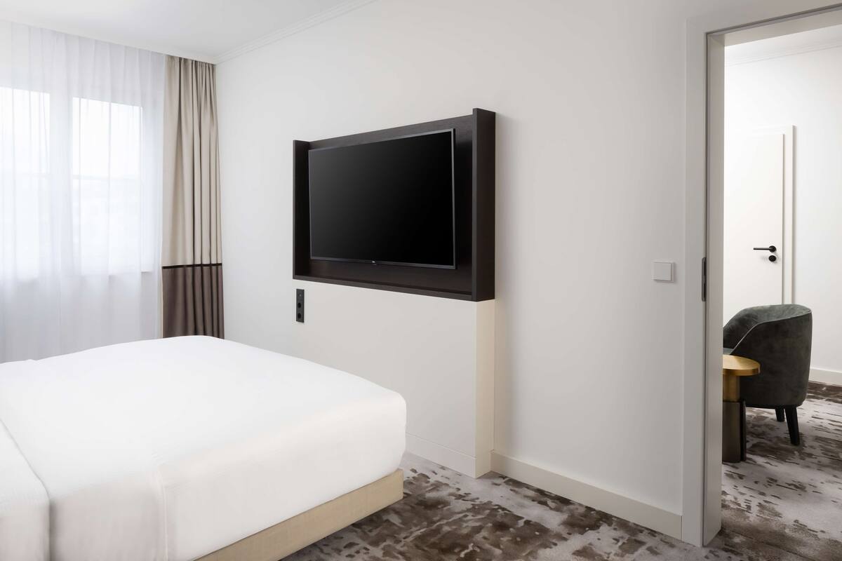 A hotel room with a bed, a mounted TV, a door leading to another room, and a chair.