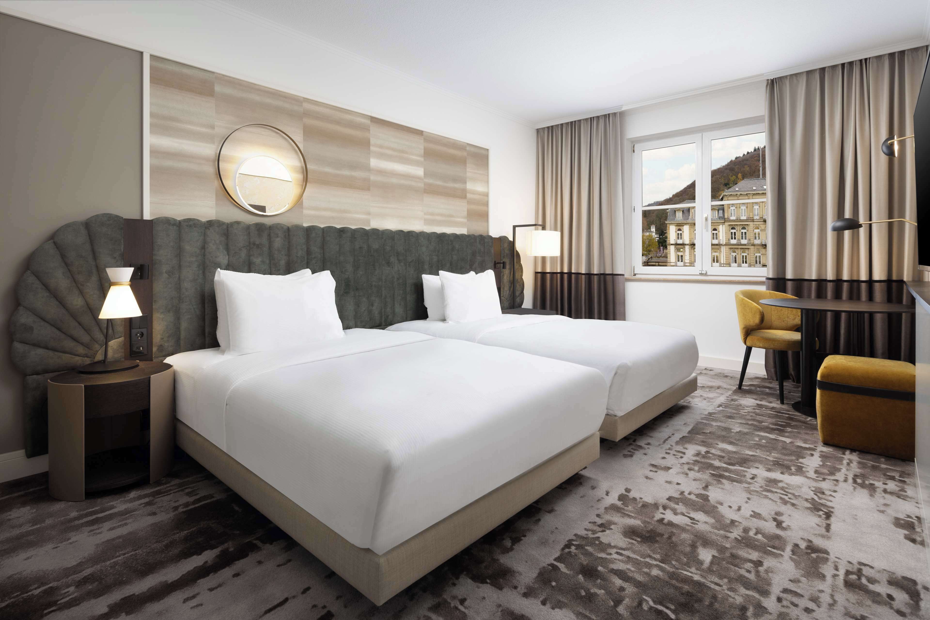 Une chambre d’hôtel avec deux lits, un bureau, une chaise et une grande fenêtre donnant sur un immeuble.