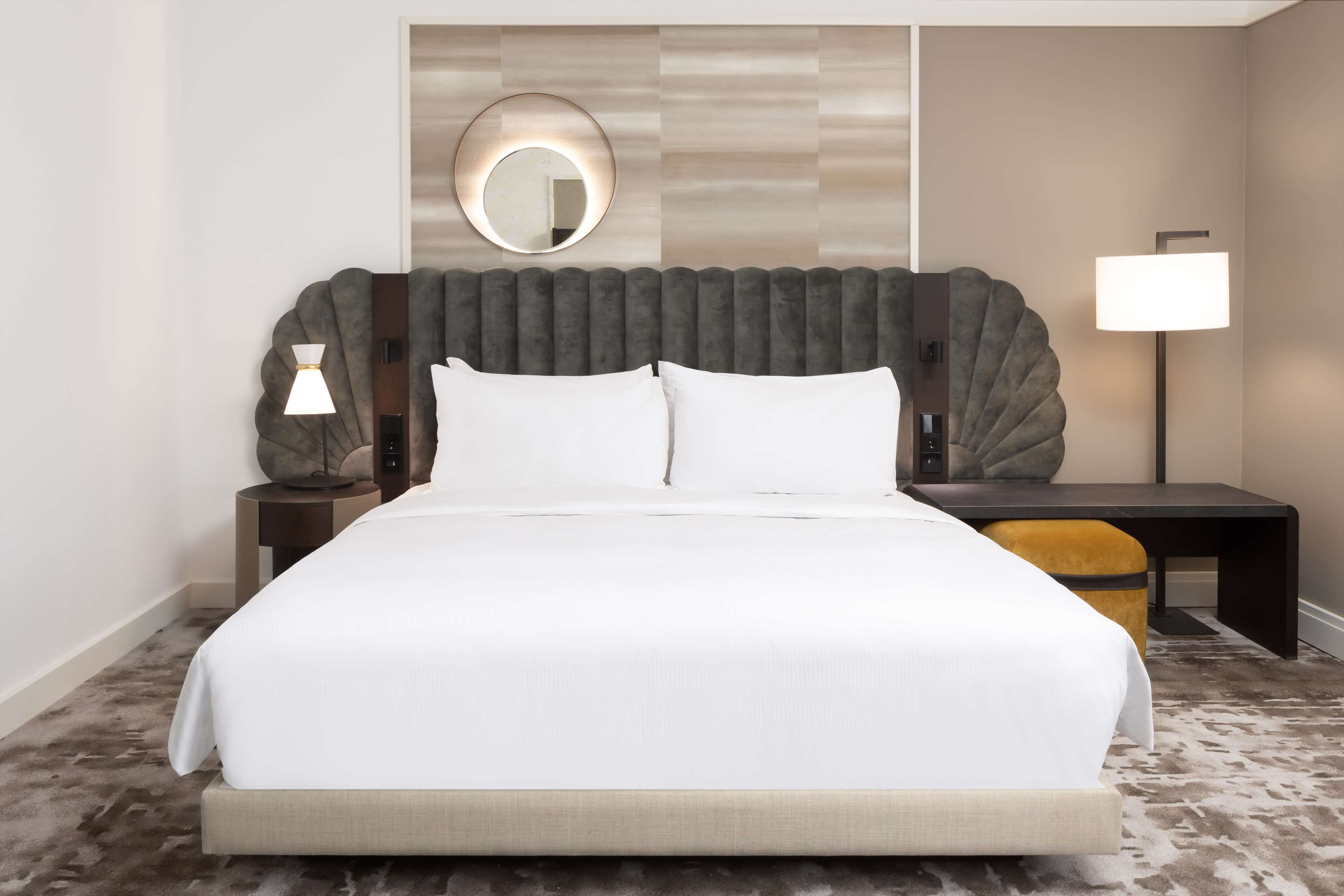 A hotel room with a large bed, a round mirror above the headboard, a bedside table with a lamp, and a small desk with a chair.