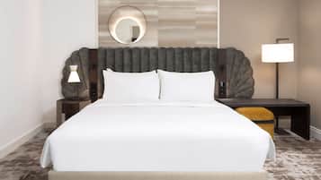 A hotel room with a large bed, a round mirror above the headboard, a bedside table with a lamp, and a small desk with a chair.