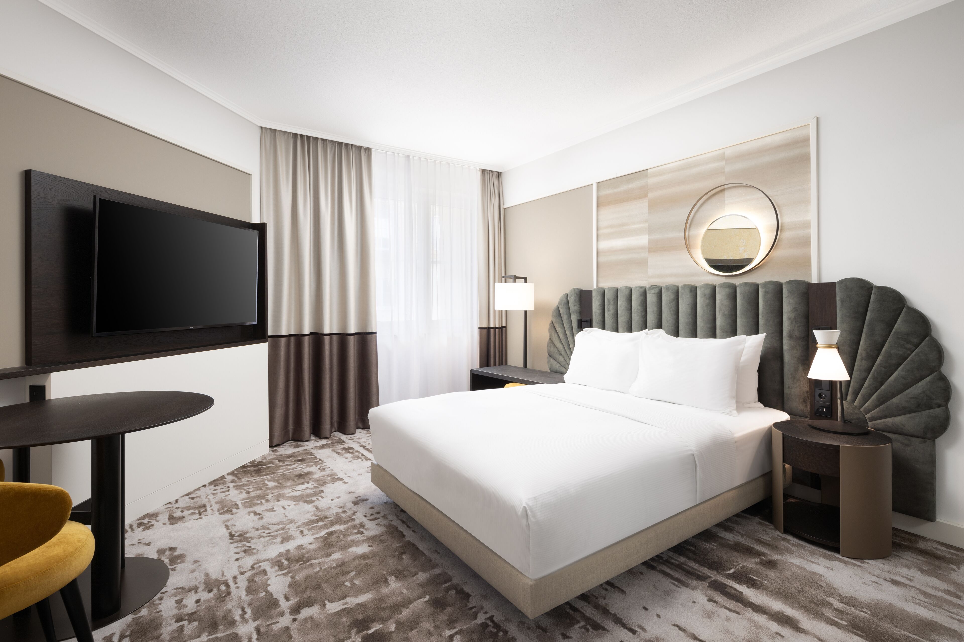 A hotel room with a large bed, a flat-screen TV mounted on the wall, a round mirror, and a small table with a lamp.