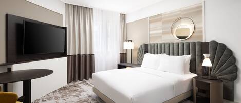 A hotel room with a large bed, a flat-screen TV mounted on the wall, a round mirror, and a small table with a lamp.