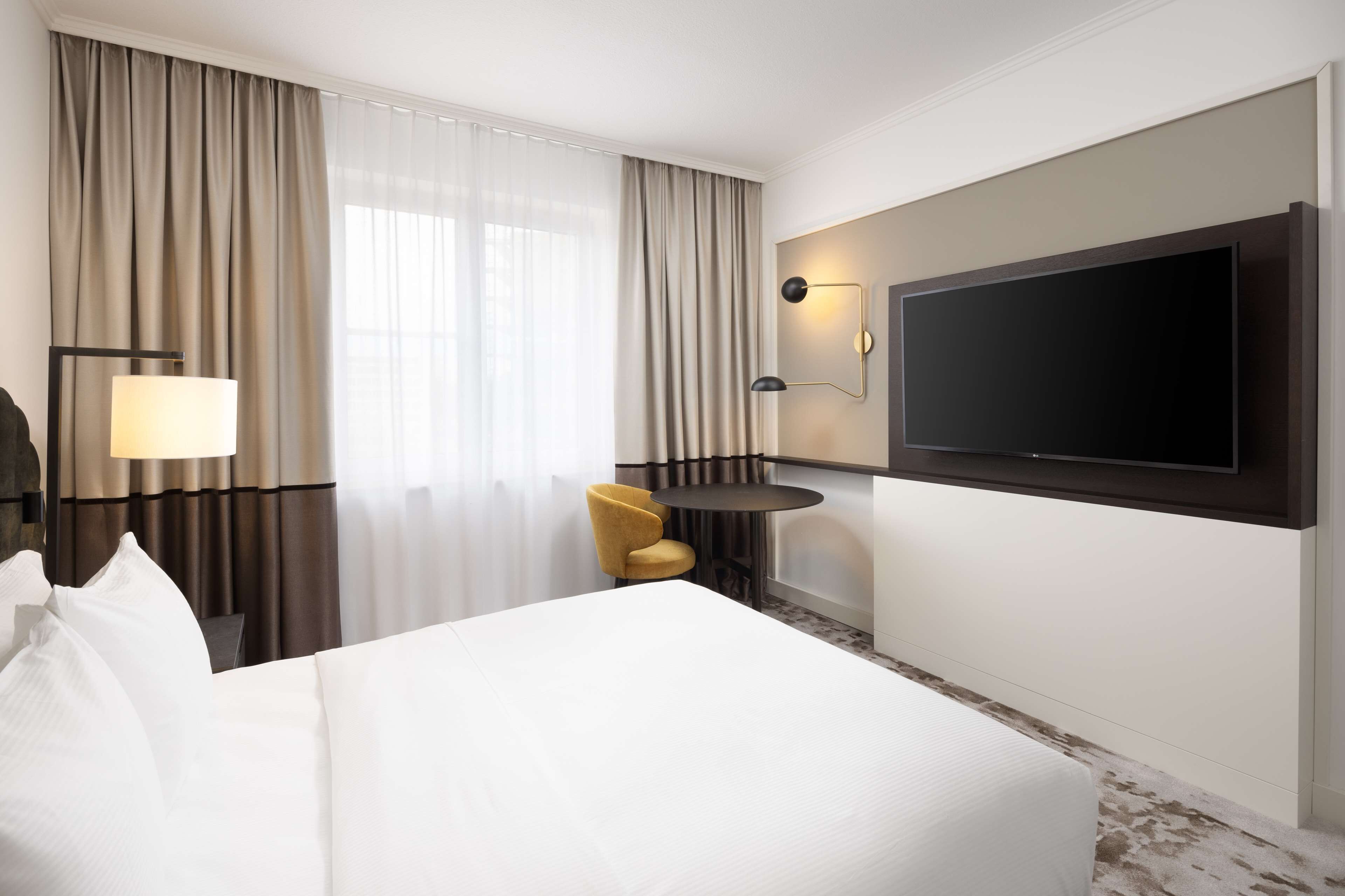 Habitación de hotel con una cama grande, un televisor de pantalla plana en la pared, una mesita redonda pequeña, un sillón amarillo y una lámpara.