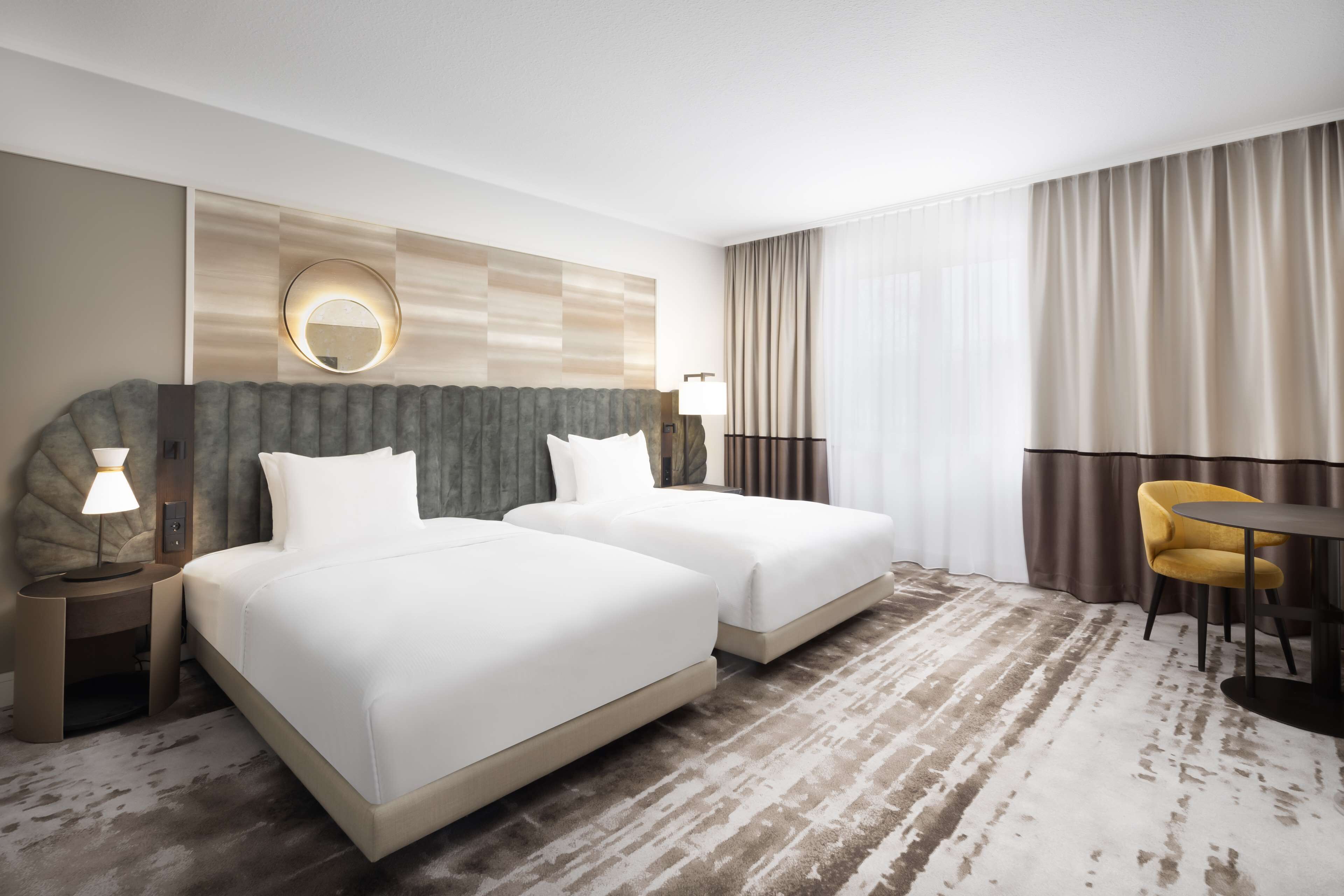 Ein Hotelzimmer mit zwei Betten, einem runden Spiegel, einem Schreibtisch mit Lampe, einem Stuhl und einem großen Fenster mit Vorhängen.